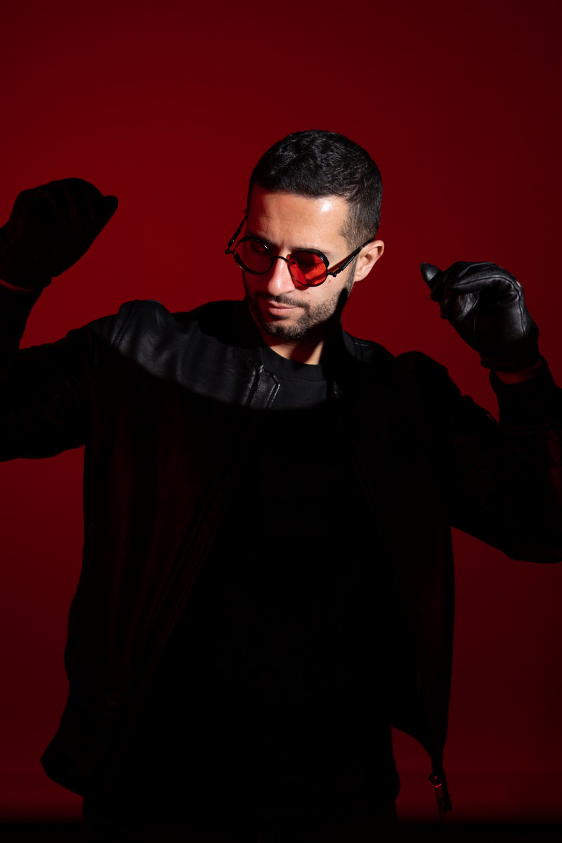 ANYMEN en portrait, lunettes teintées et veste noire, sur fond rouge